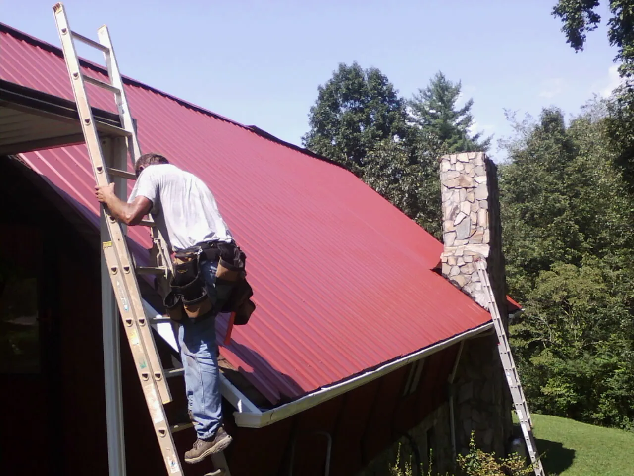 Roofer on a ladder beside a red metal roof and stone chimney — Soffit & Fascia Repair by Roofing Brothers in Wakonda